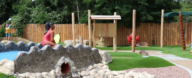 Adventure-Golf-Anlage mit Wellenfigur, künstlichem Rasen, Felsen und bunten Themen-Elementen wie Palme, Krabbe und Sandburg.